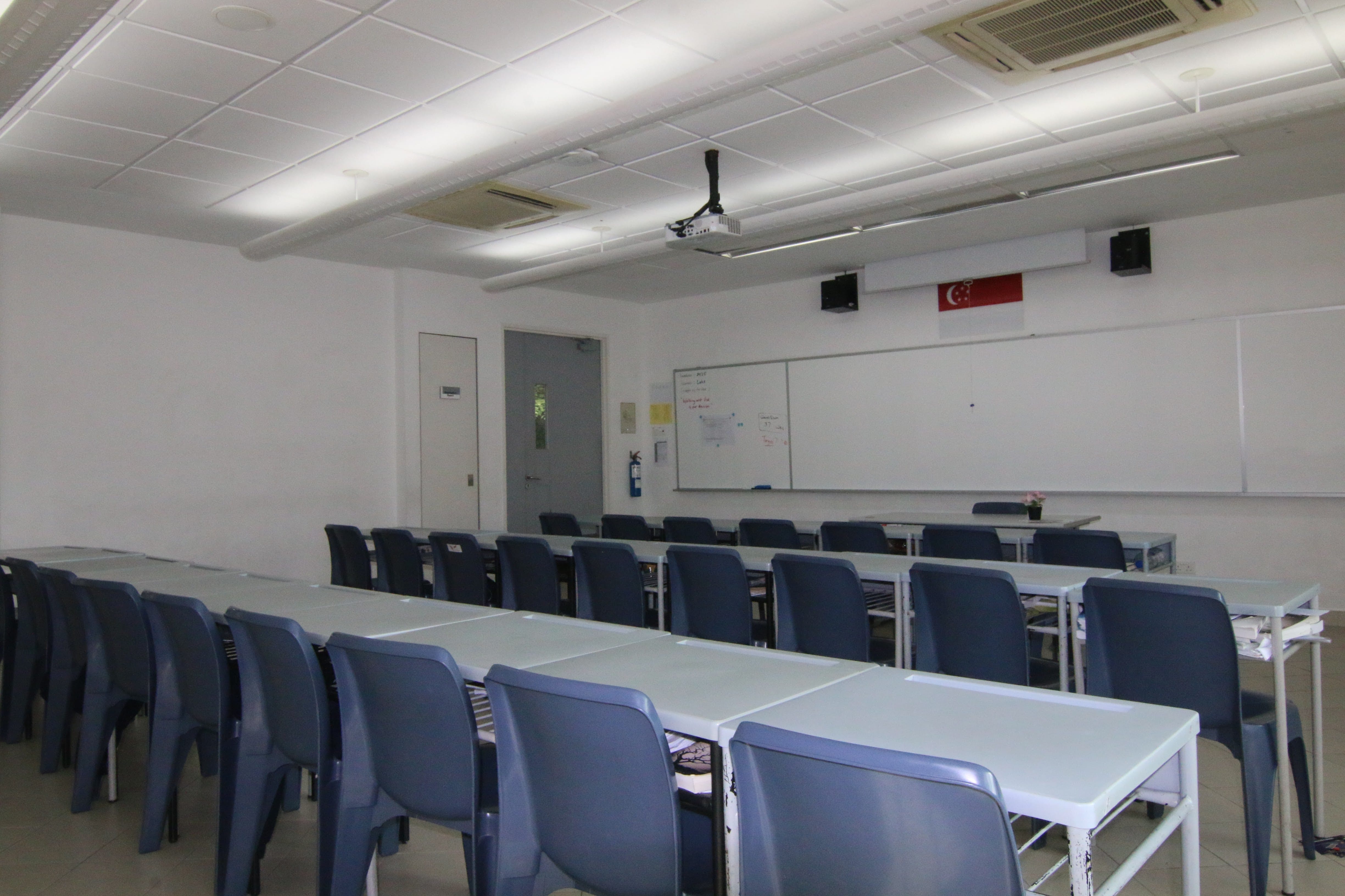 Empty classroom with rows of blue chairs and long tables facing a white board at the front.